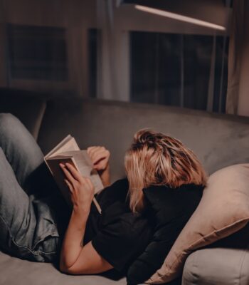 lit Girl reads book in the evening on couch. Night lamp light. Relaxing evening. Large window. Modern living room. Velvet pillows. Night light. Blonde woman reads fiction book. Quarantine stay at home.